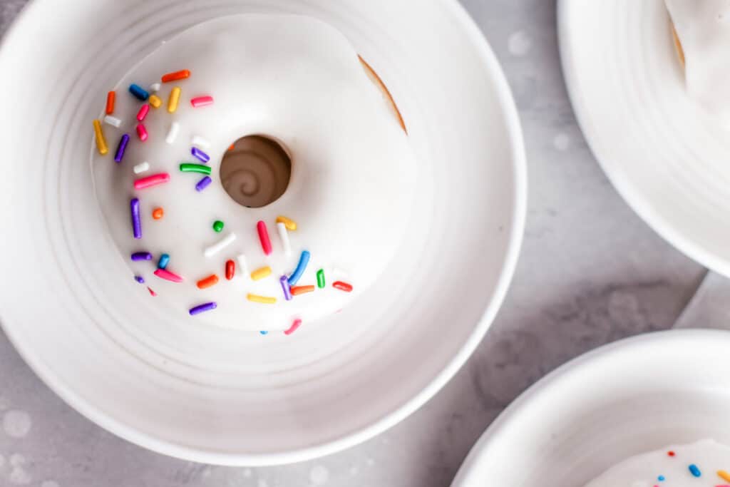 one baked vanilla cake donut with white frosting and colored sprinkles on a white plate