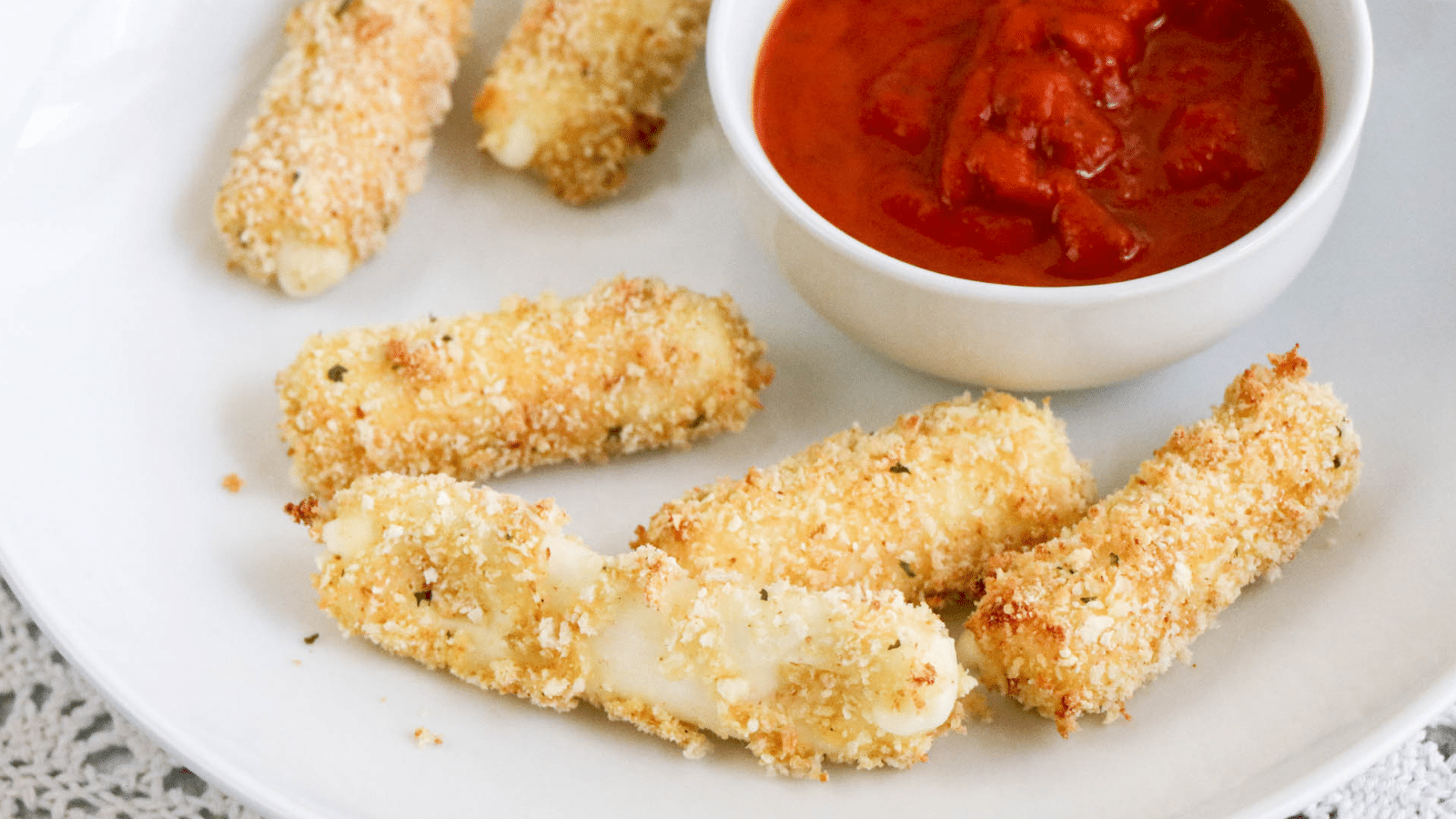 air fryer mozzarella sticks on a white plate with one stretched out showing the cheese inside and marinara sauce