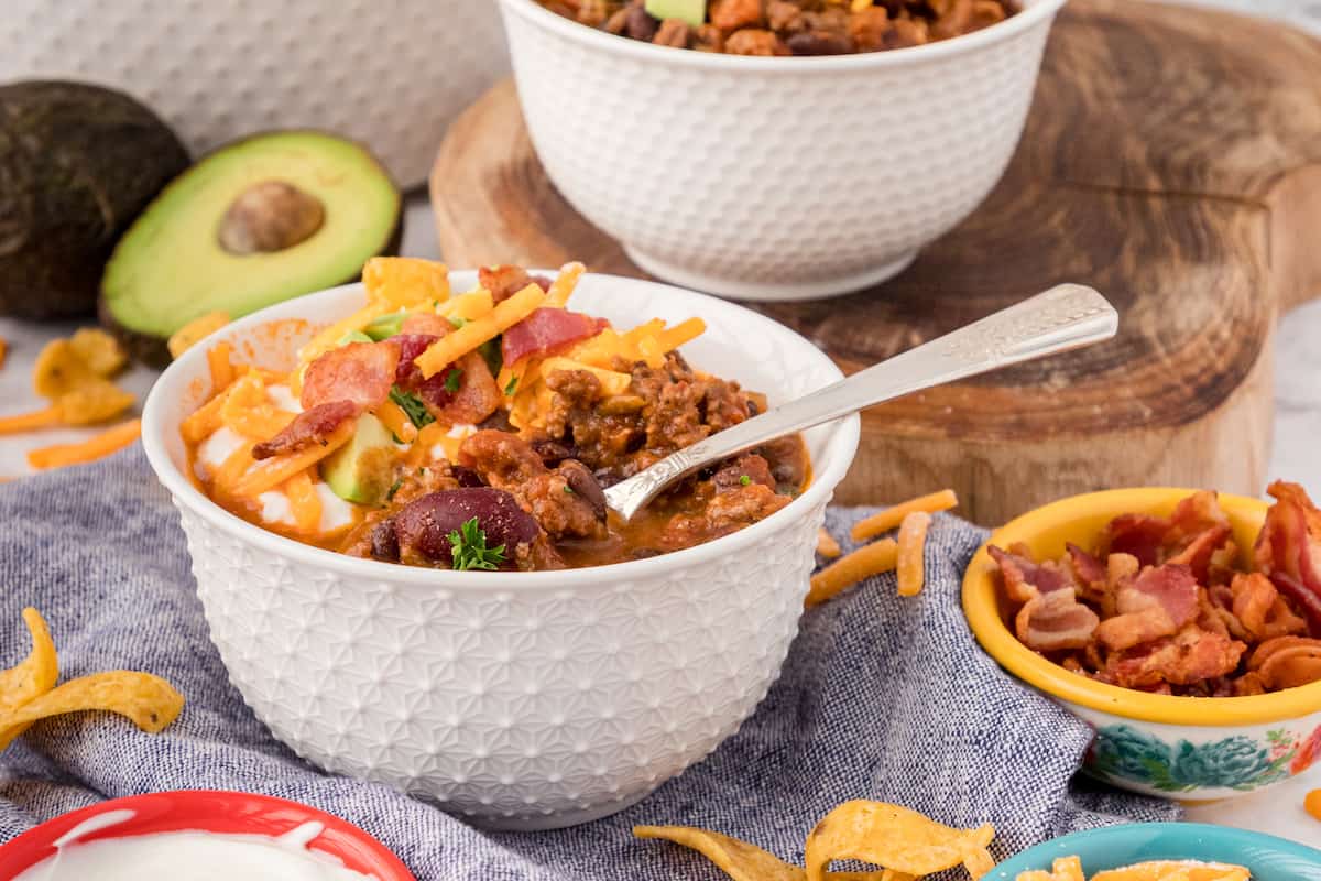 world's best chili with bacon in a white bowl topped with crumbled bacon, cheddar cheese, and chopped avocado