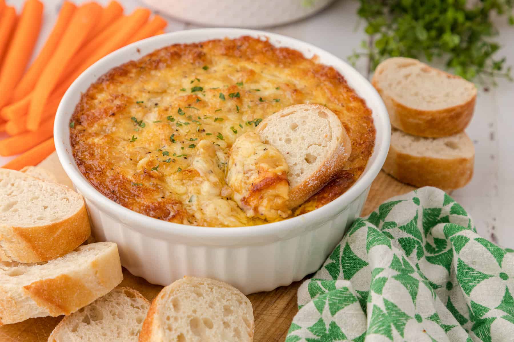hot onion dip with swiss cheese in a white baking dish, surrounded by crostini and carrot sticks