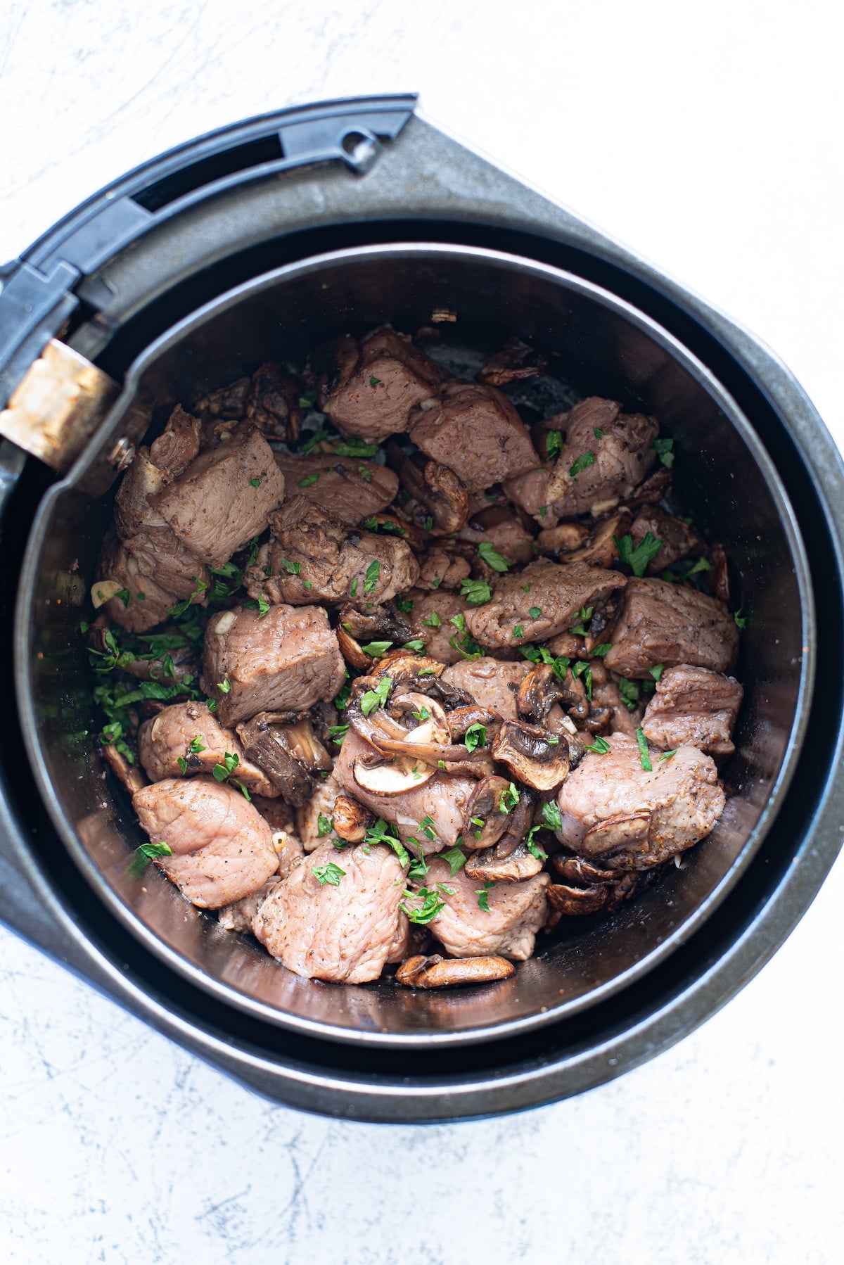 air fryer steak bites with mushrooms in the air fryer basket, garnished with chopped parsley