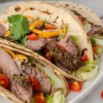 a close up of three marinated flank steak tacos with toppings on a white plate