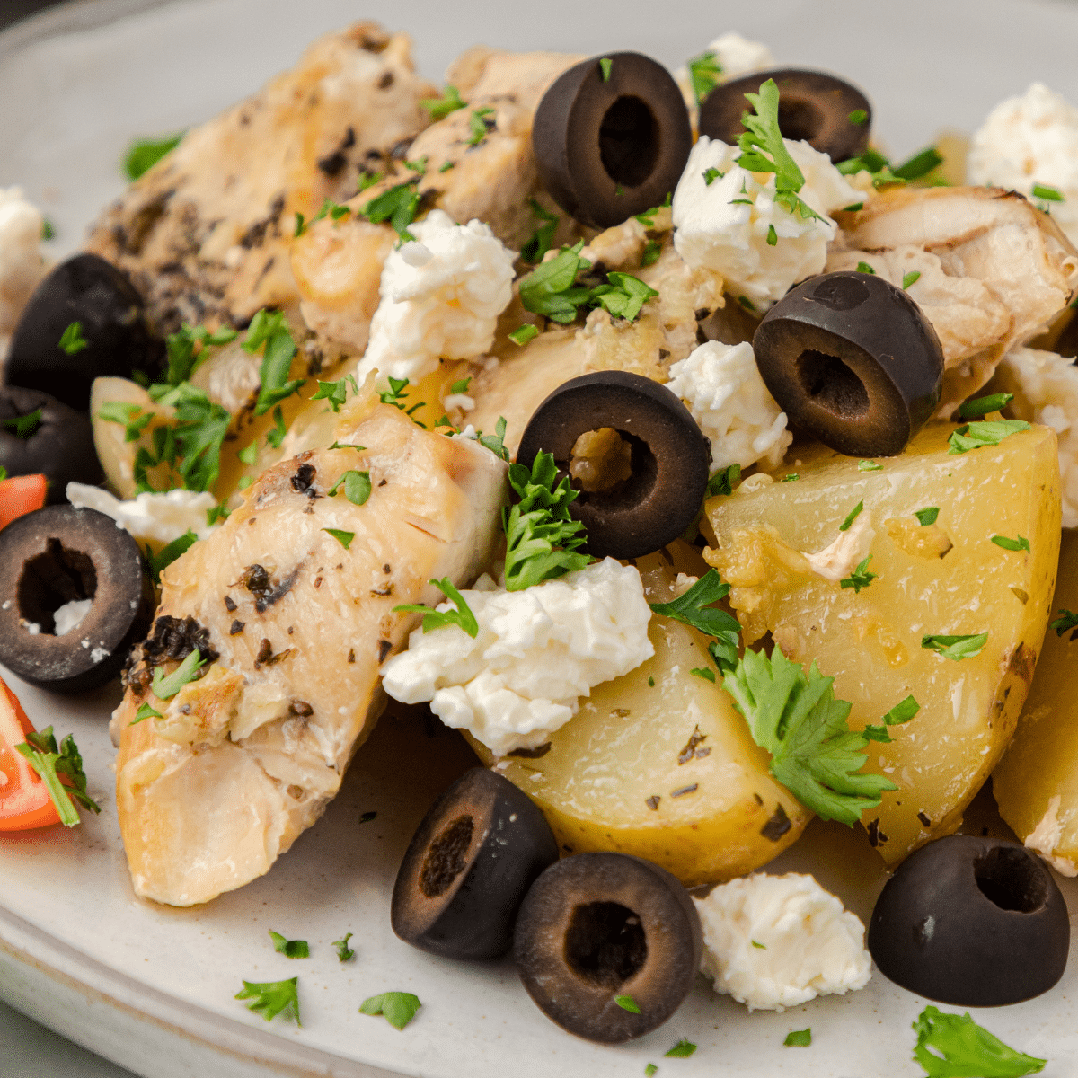 CrockPot chicken and potatoes on a white plate topped with black olives, feta cheese, and chopped parsley