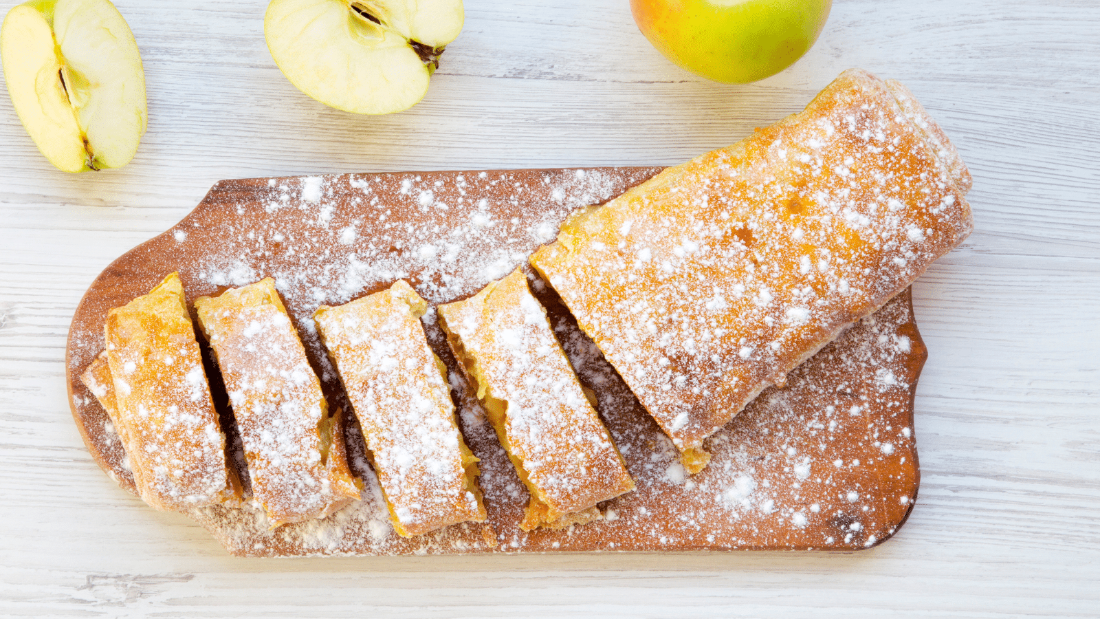 a partially sliced Caramel Apple Strudel made with puff pastry on a wooden cutting board with an apple