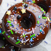 a close up of a baked chocolate donut with chocolate ganache glaze and multicolored sprinkles