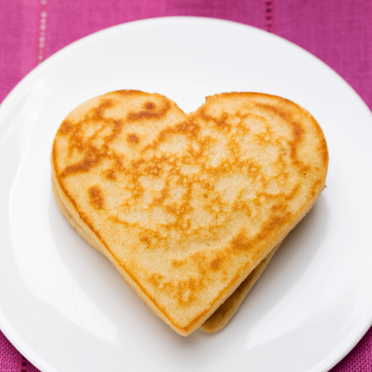 two heart-shaped pancakes on a white plate on top of a pink tablecloth