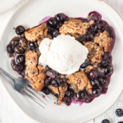 one serving of old-fashioned blueberry cobbler with a scoop of vanilla ice cream on a white plate with a fork