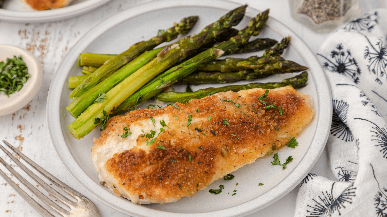 one baked parmesan crusted chicken breast with asparagus on a white plate