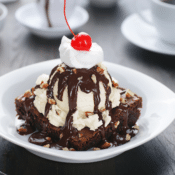 gluten-free hot fudge brownie sundae with vanilla ice cream, whipped cream, chopped nuts, and a cherry in a white bowl