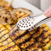 picking up one piece of grilled zucchini which is on a platter with grilled summer squash