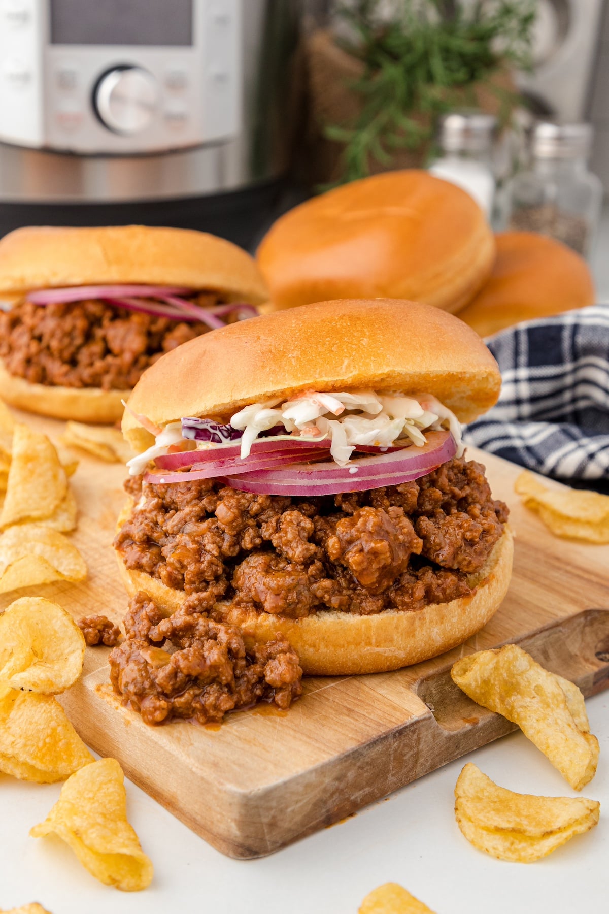 an Instant Pot sloppy joe sandwich with slices of red onion and coleslaw on top surrounded by potato chips and the Instant Pot in the background