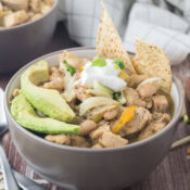 a close up of a bowl of green chili chicken soup topped with avocado slices, shredded cheddar cheese, sour cream, green onions, and tortilla chips