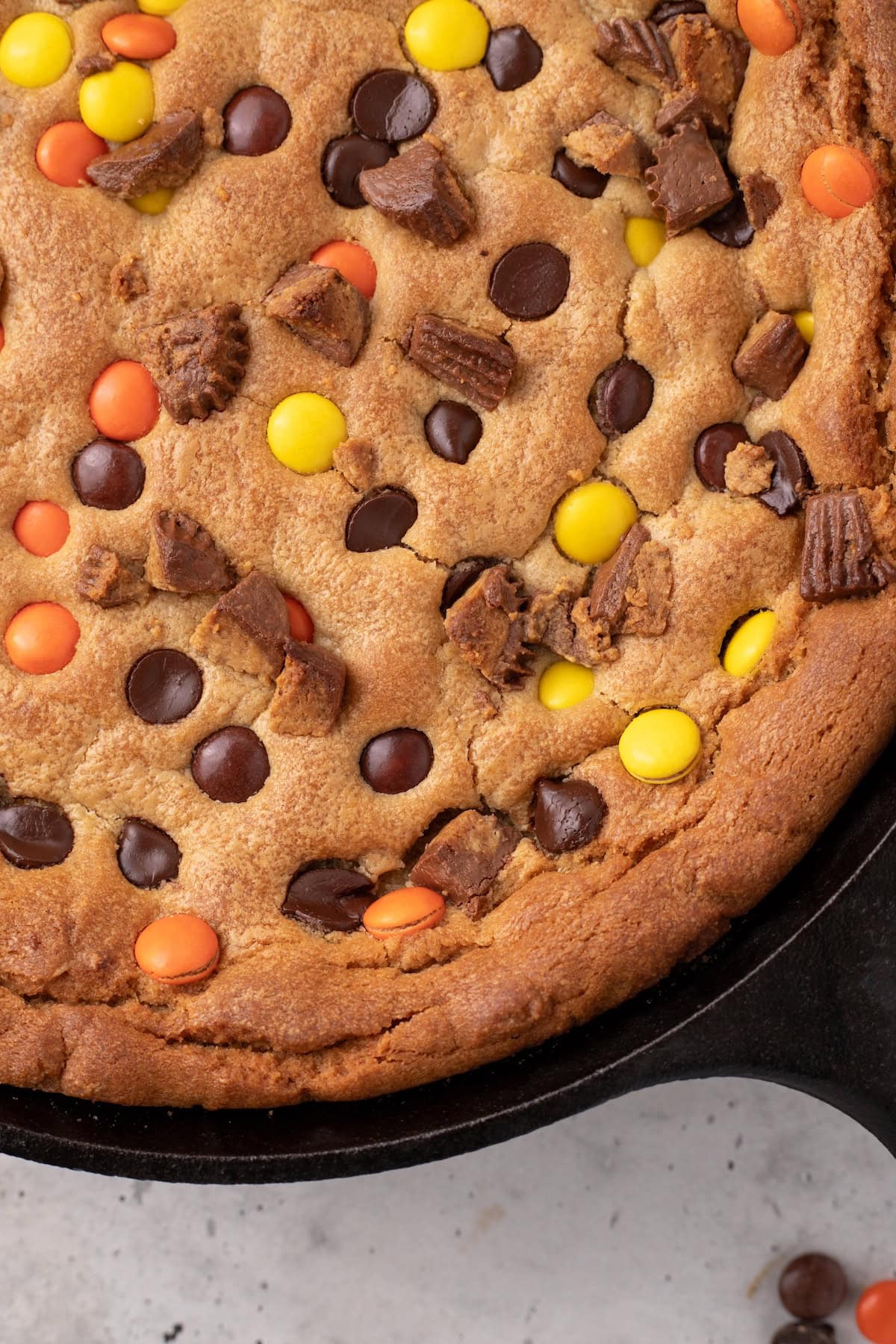 a close up of the ultimate peanut butter skillet cookie showing PB cups, Reese's Pieces, and chocolate chips on top of a golden brown peanut butter cookie cake made in a cast iron skillet