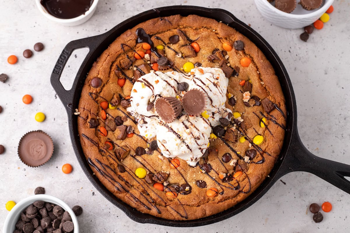 the ultimate peanut butter skillet cookie showing PB cups, Reese's Pieces, and chocolate chips on top of a golden brown peanut butter cookie cake topped with vanilla ice cream and hot fudge