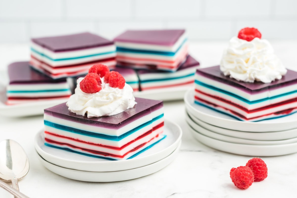 many large squares of red white and blue layered jello topped with whipped cream and fresh red raspberries