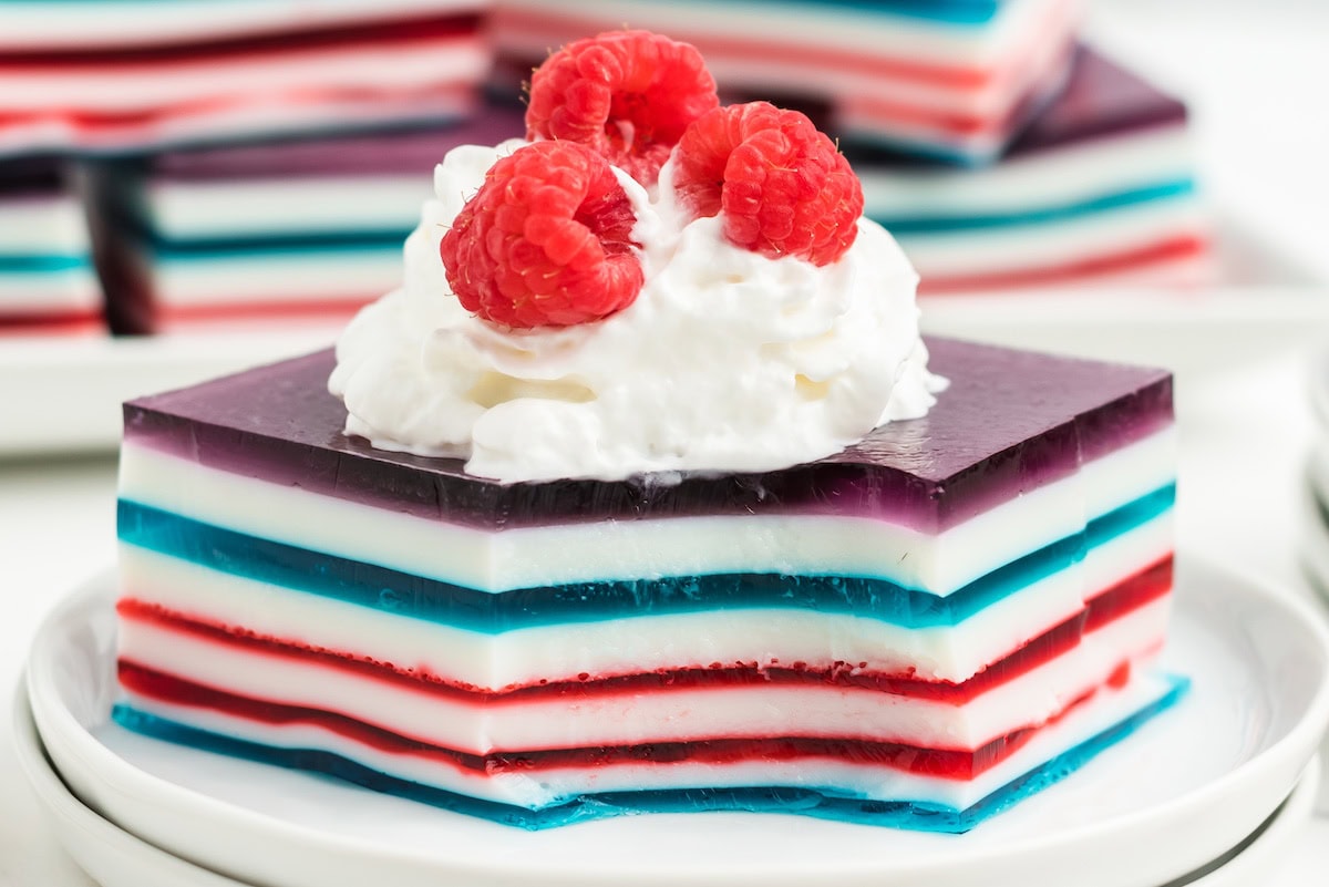 a large square of red white and blue layered jello topped with whipped cream and fresh red raspberries with one bite removed