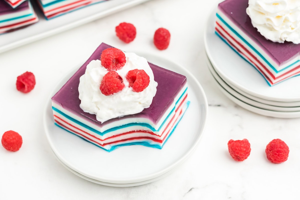 two large squares of red white and blue layered jello topped with whipped cream and fresh red raspberries on white plates
