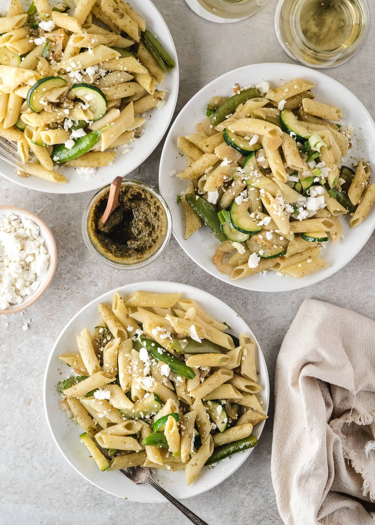 three servings of easy pasta primavera on white plates with pesto and olive oil