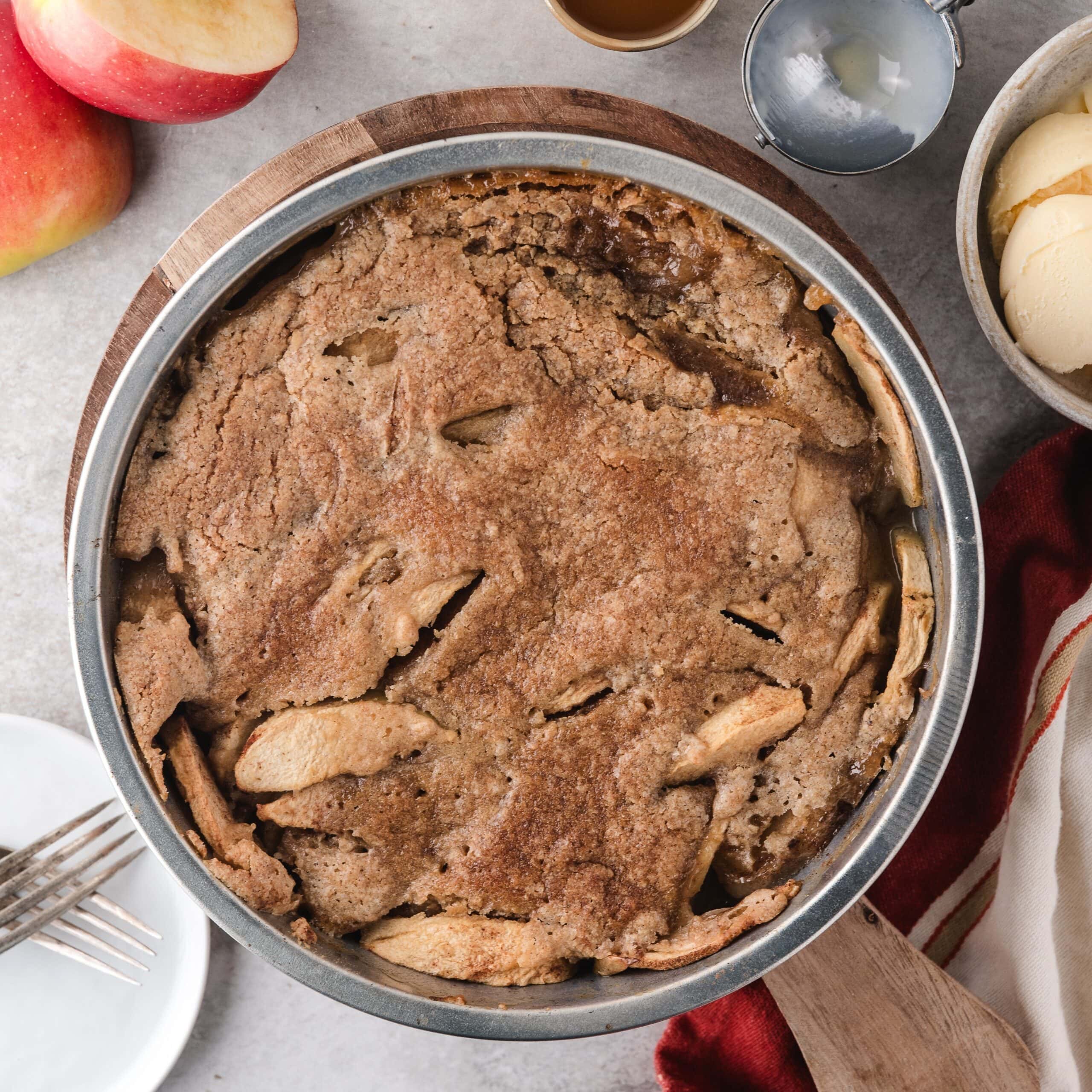 Best Apple Crisp without Oats right from the oven. The whole healthy crisp is in a 9x9 circular pan. Around the pan is added elements as a tea towel, ice cream scoop, an apple, forks and plates and a bowl of vanilla ice cream.