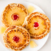 three golden brown pineapple upside down pancakes on a white plate surrounded by pineapple chunks