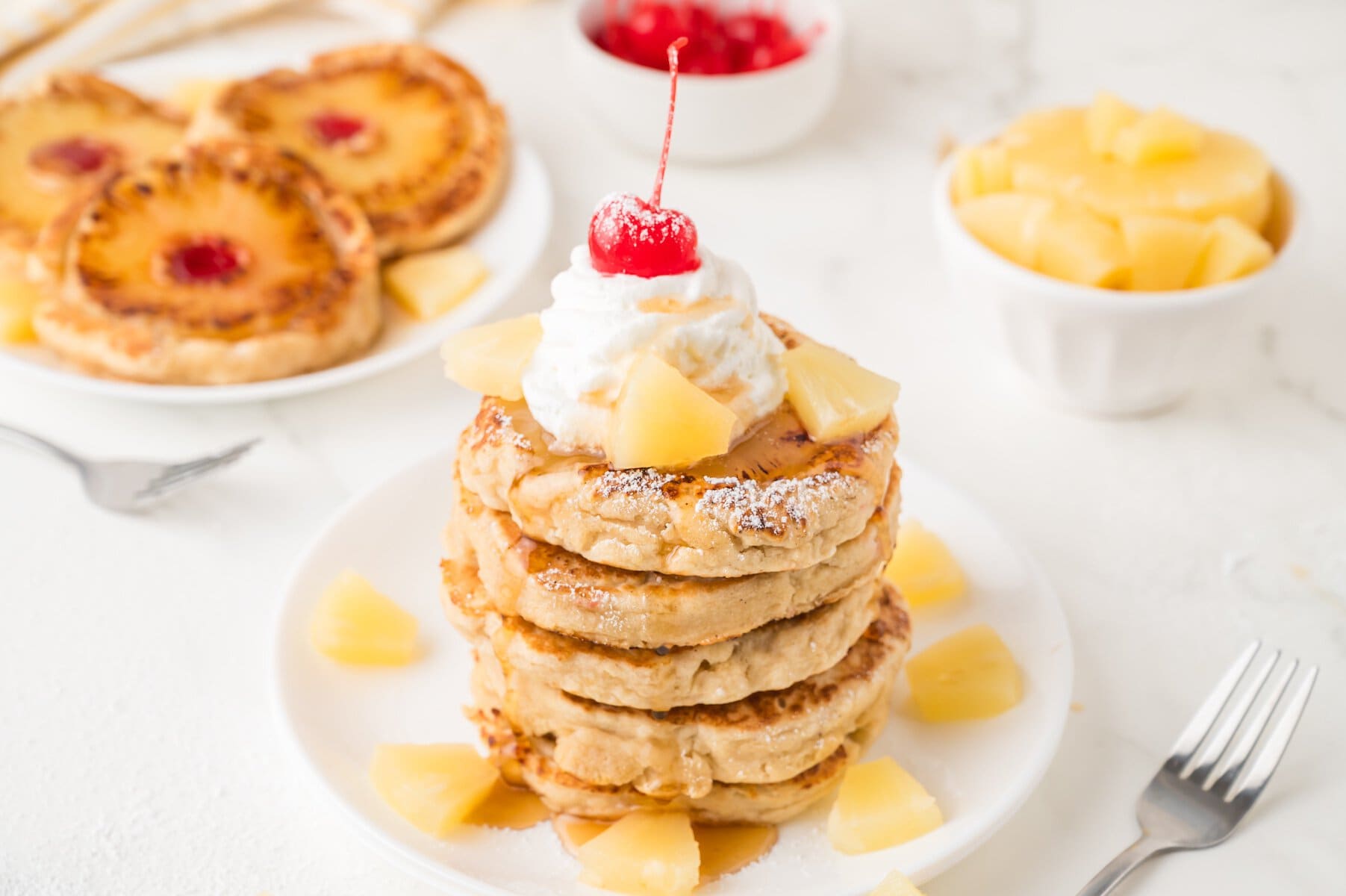 a stack of pineapple upside down pancakes with pineapple chunks, whipped cream, and a cherry