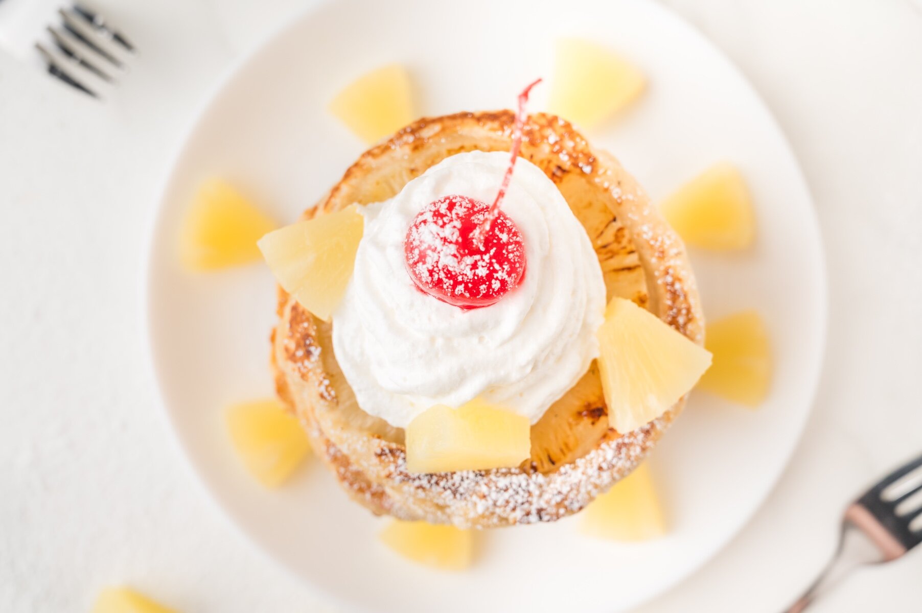 a stack of pineapple upside down pancakes with whipped cream and a cherry and dusted with powdered sugar and surrounded by pineapple chunks