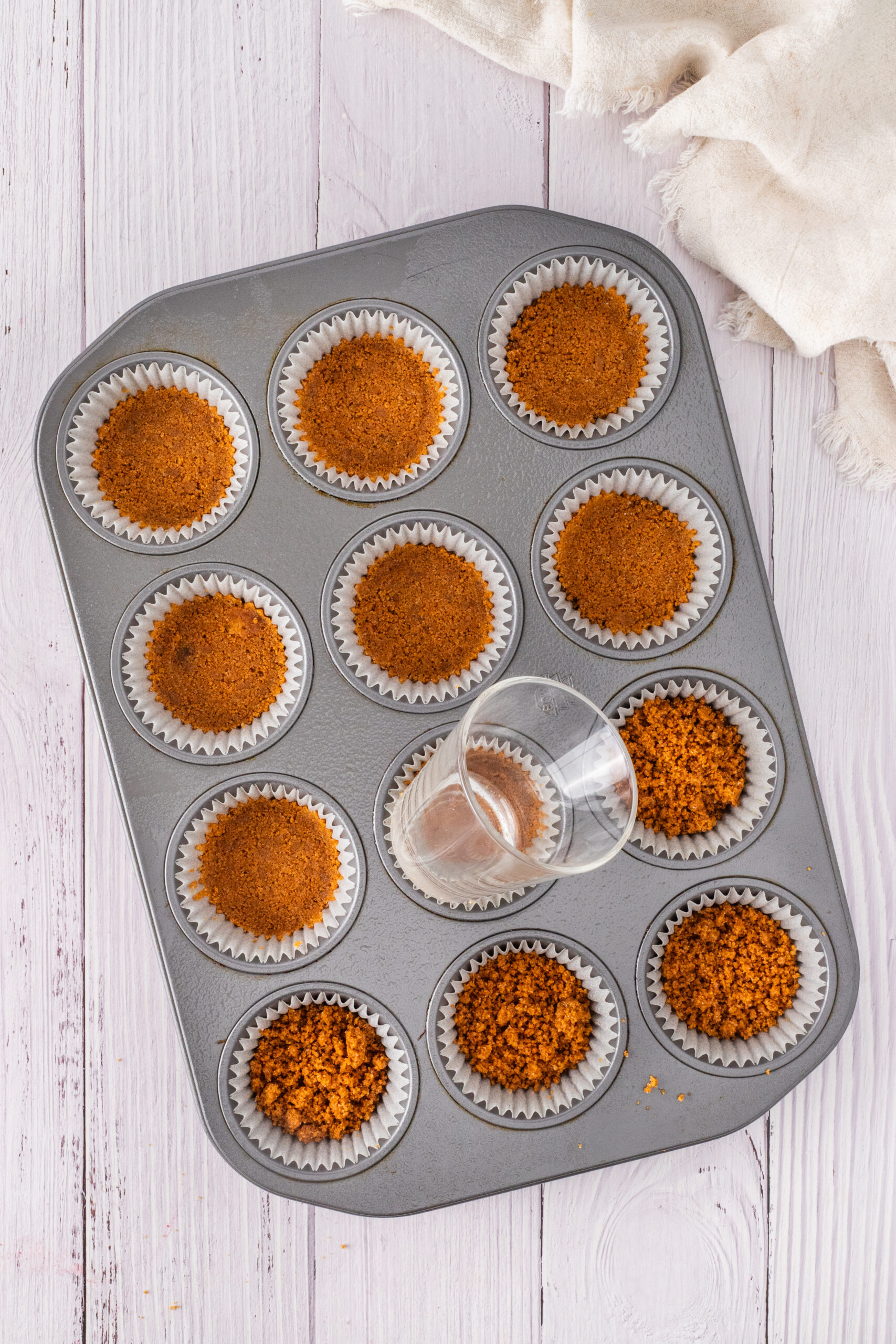 The graham cracker bottom layer is being added to the muffin cups & pressed down to finish the crust