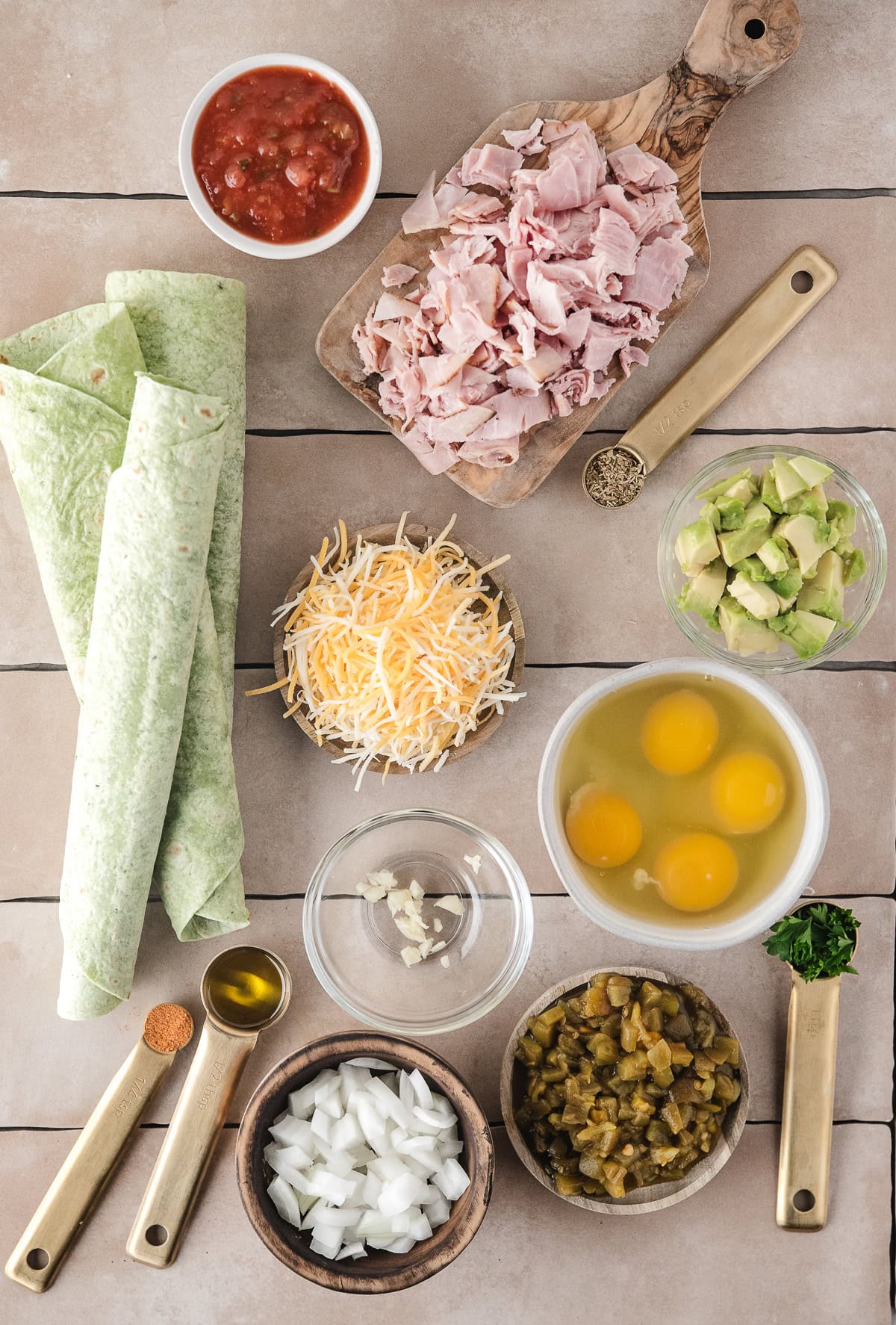 All ingredients pictured: onion, green chilis, eggs, ham, salsa, avocado, shredded cheese, tortillas, spices and fresh parsley