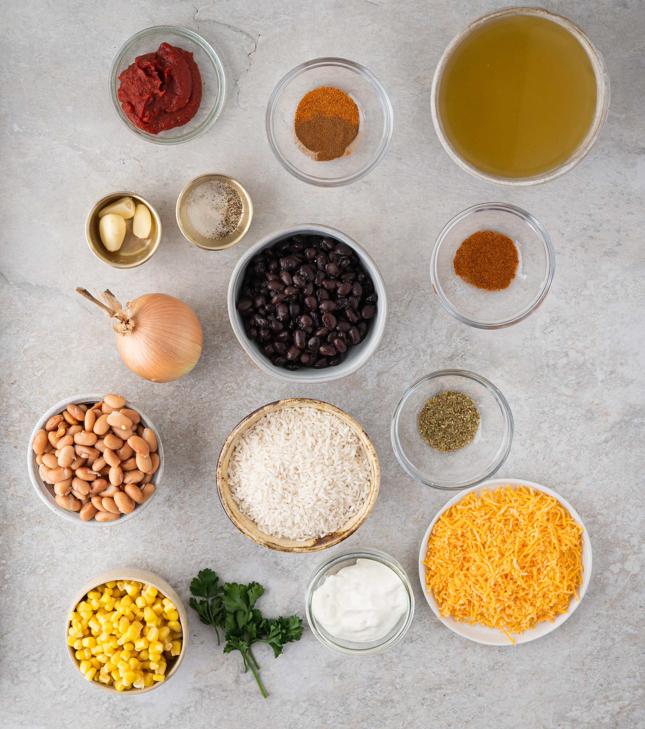 All ingredients pictured to make Mexican Rice and Beans: corn, cheddar cheese, sour cream, fresh parsley, pinto beans, long grain white rice, garlic, salt and pepper, black beans, onions, chicken broth and seasonings