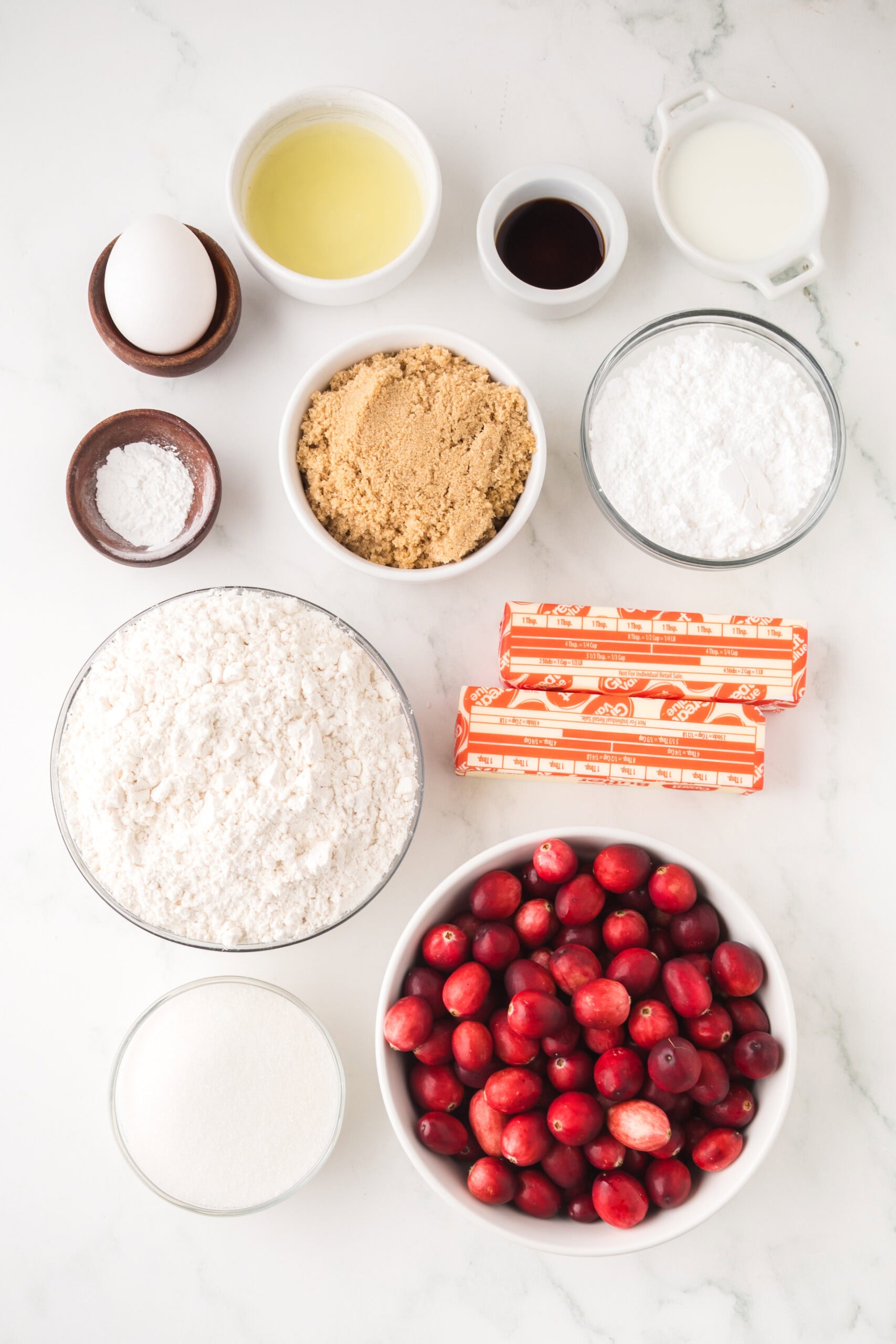 All ingredients to create cranberry bars pictured: fresh cranberries, sugar, lemon juice, butter. brown sugar, an egg, powdered sugar, vanilla extract, baking soda, baking powder, flour, whole milk, granulated sugar