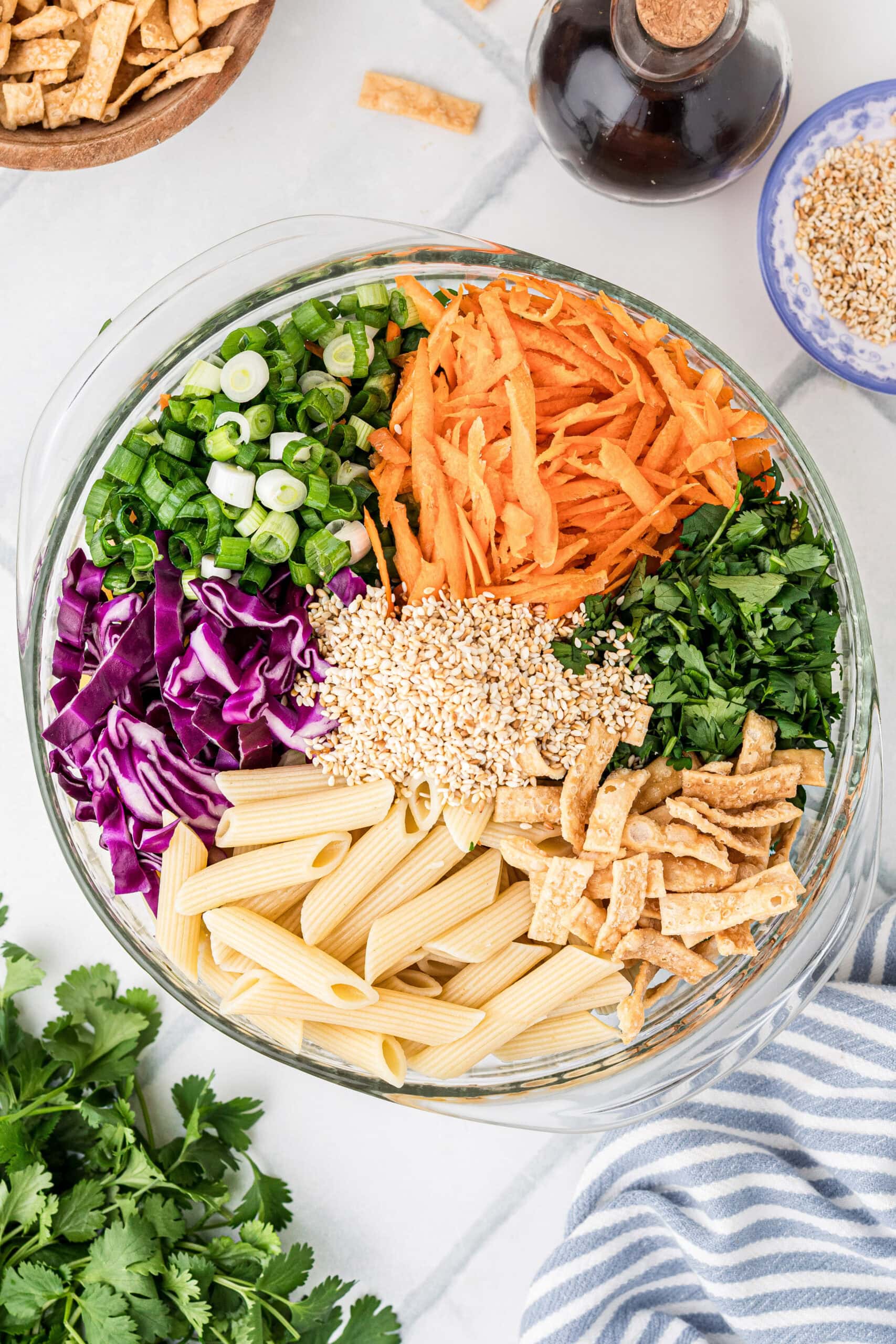 Process of putting the salad together: adding red cabbage, carrots, green onion, fried wontons, toasted sesame seeds, and fresh cilantro over the cooked pasta salad.