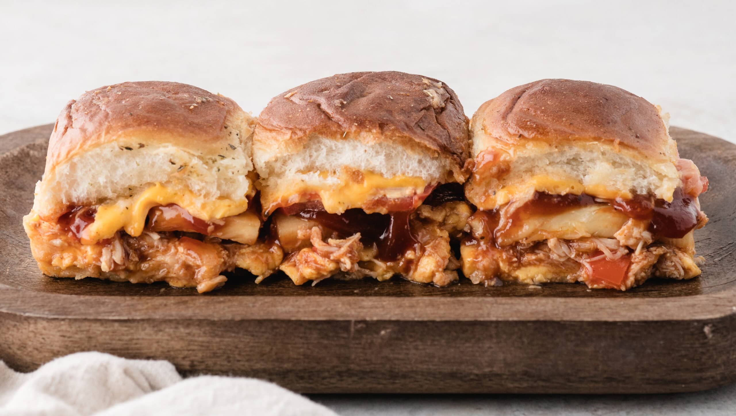 Three BBQ sliders on a wooden tray, showing its texture and layers of all ingredients