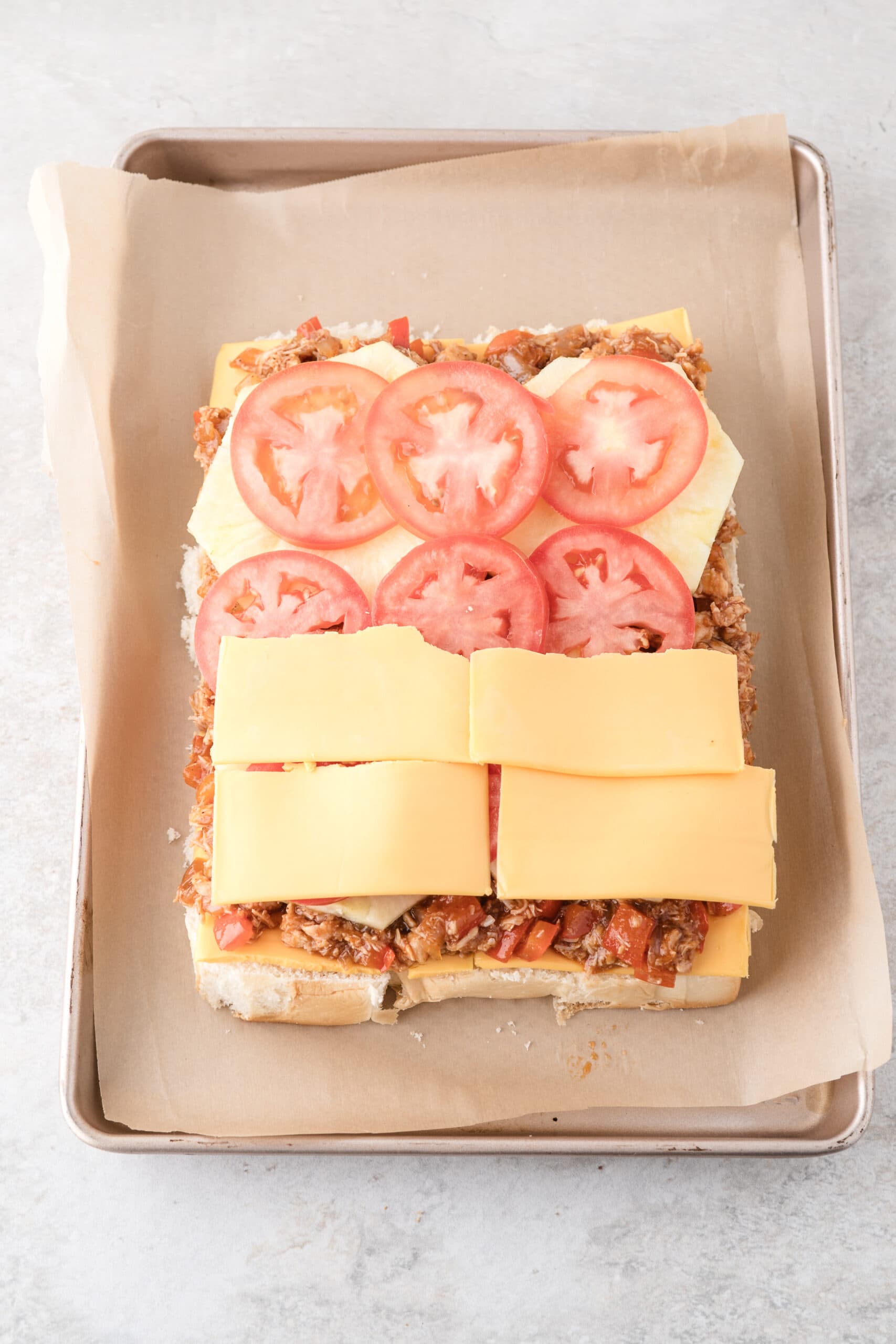 Adding cheese on top of tomatoes, pineapple, and bbq chicken on the bread bottom of the buns