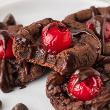 several chocolate covered cherry cookies topped with a maraschino cherry and drizzled with cherry chocolate ganache, and one has a bite out to show the fudgy chocolate texture inside