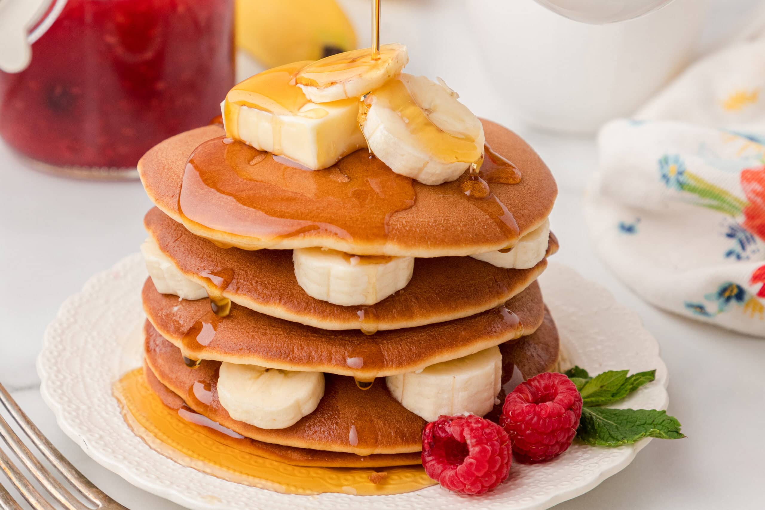 Layers of banana pancakes, with bananas in the middle, syrup is being poured on top of the stack