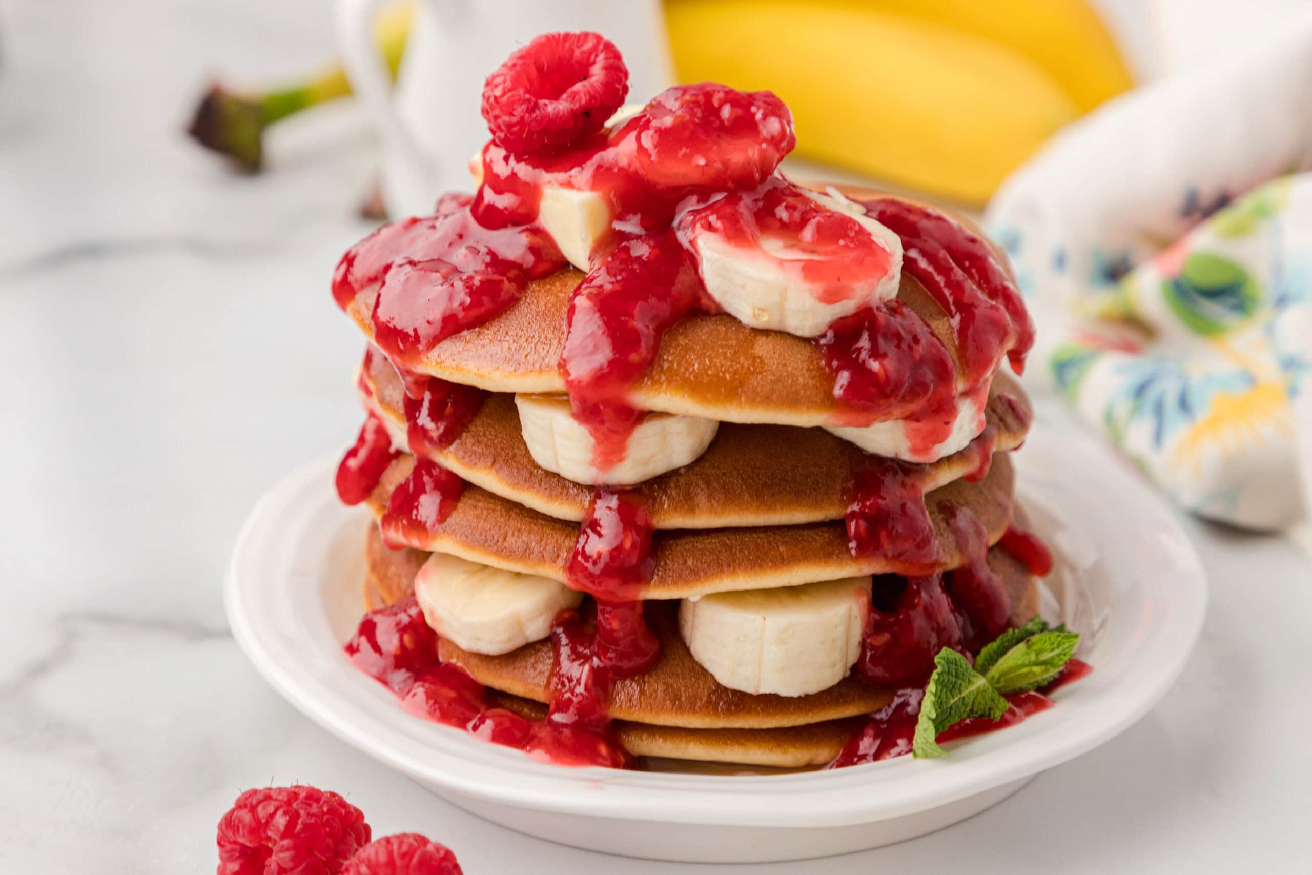 Raspberry sauce is poured on top of banana protein pancakes, with pancakes in layers with bananas in the middle of the layers.