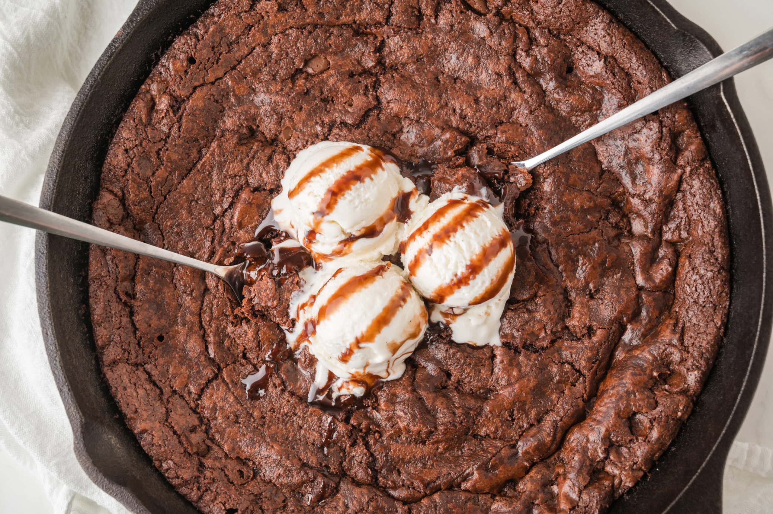 Brownie skillet all baked, topped with vanilla ice cream and chocolate sauce, with two spoons