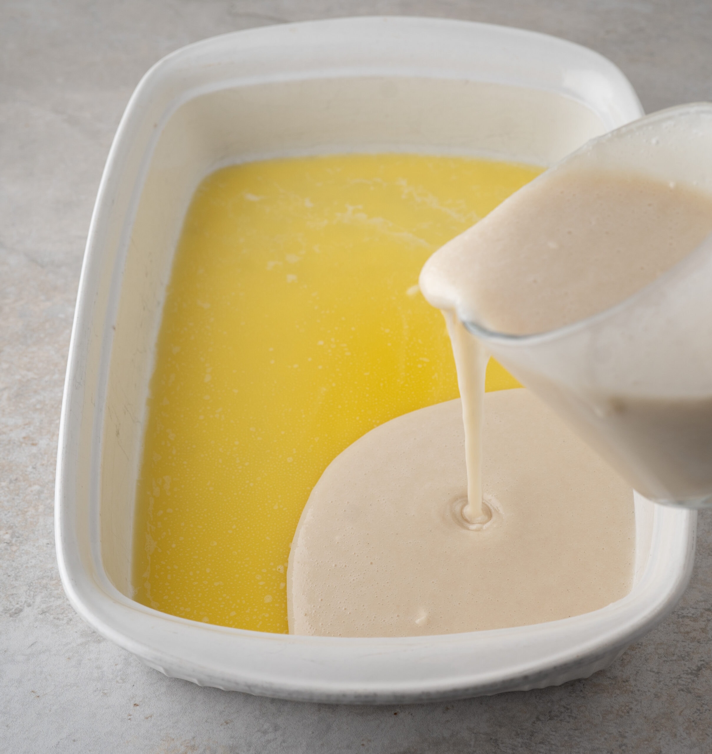 Batter created from flour, sugar salt and baking powder and vanilla bean paste is being poured on top of melted butter in baking dish