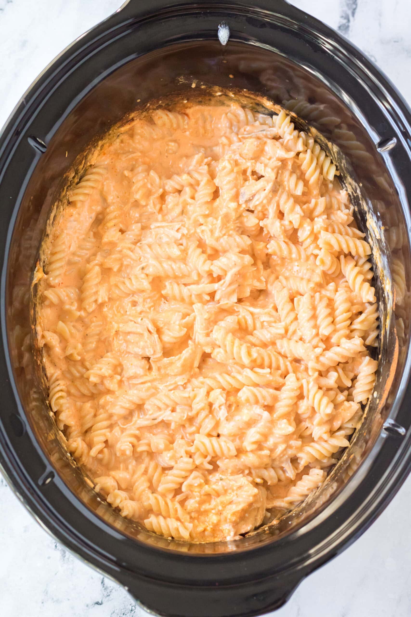 Pasta cooked and drained and then being added to the buffalo chicken mixture in the crockpot.