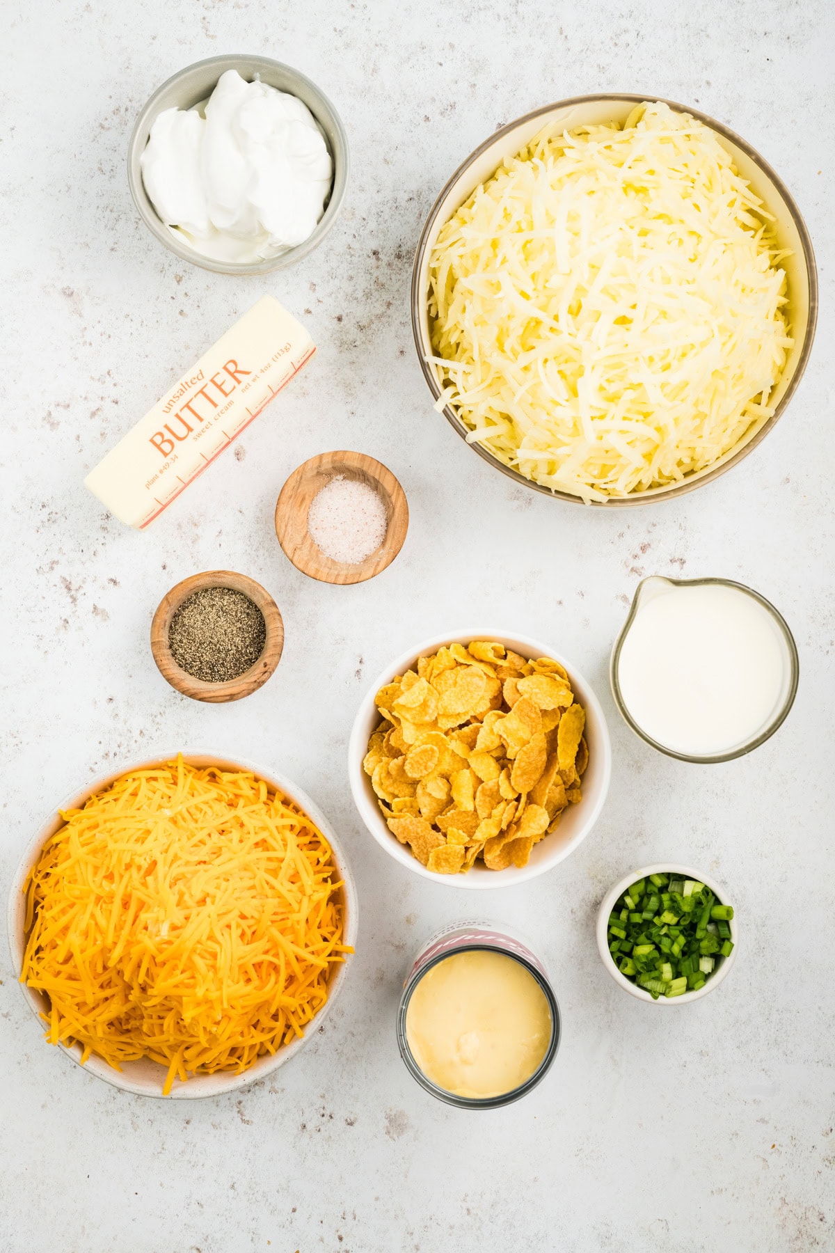 All ingredients pictured to create cheesy potatoes: cheddar cheese, melted butter, hash browns, salt, pepper, corn flakes, sour cream, milk, cream of chicken soup and green onion