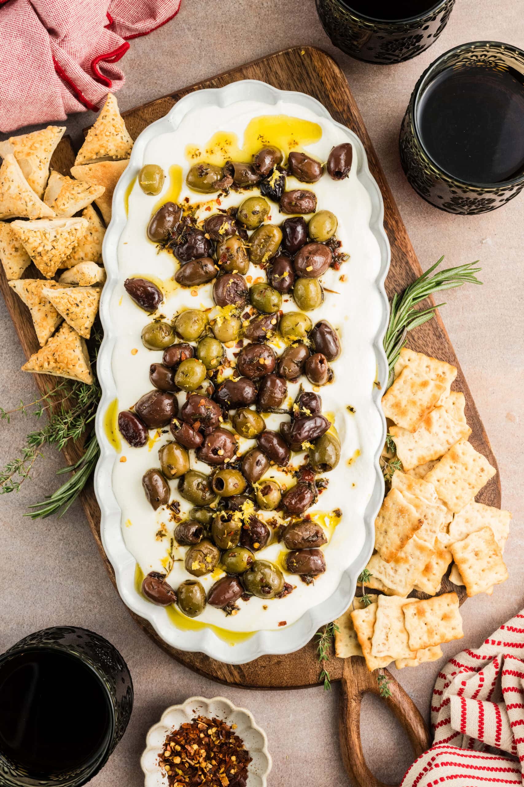 Whipped Feta dip with olives on a platter, theres crackers surrounding it with wine, red pepper flakes,