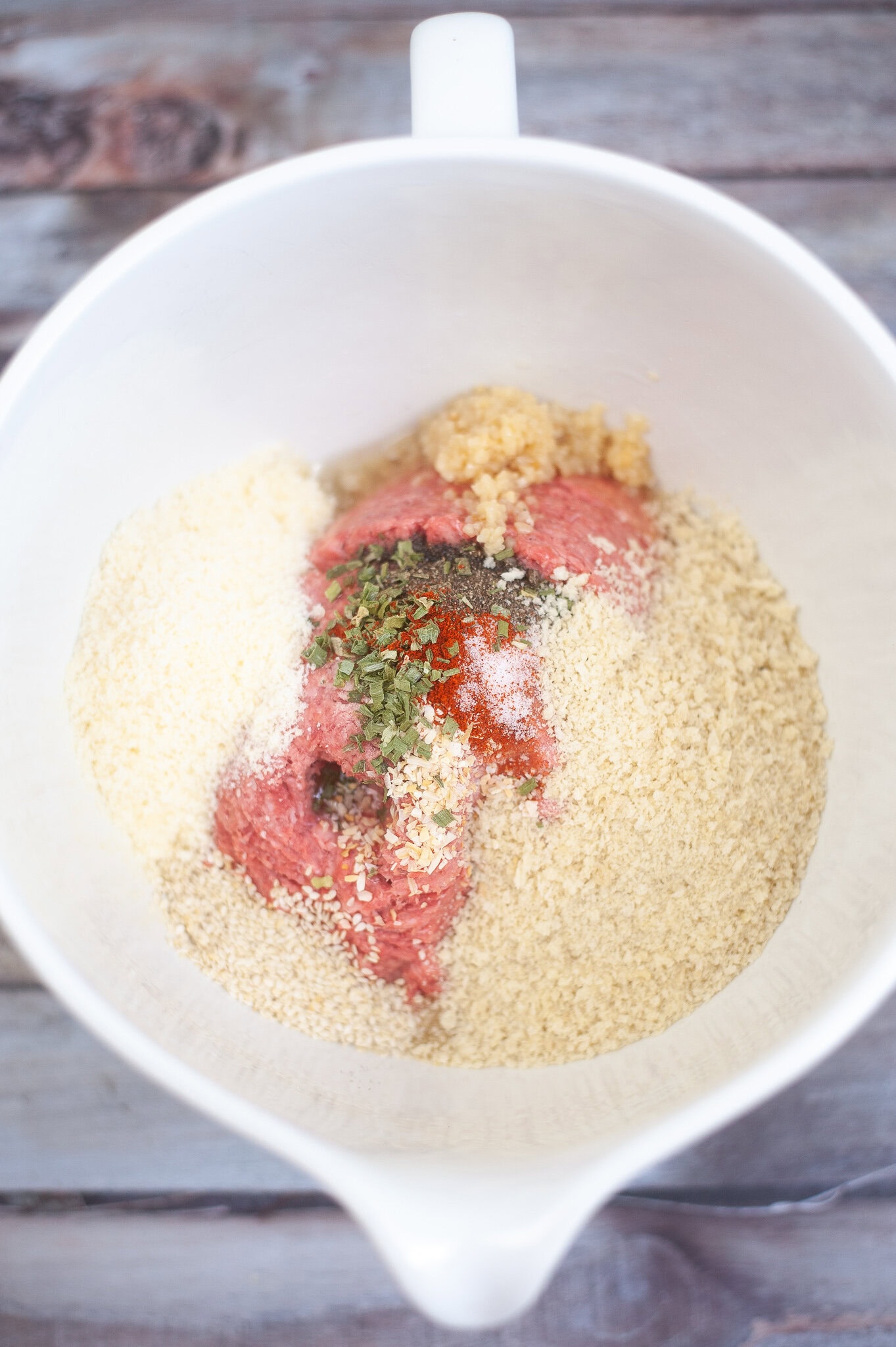 In a mixing bowl there is ground beef, salt, pepper, onion powder, paprika, eggs, panko breadcrumbs, parmesan cheese, sesame seeds and parsley