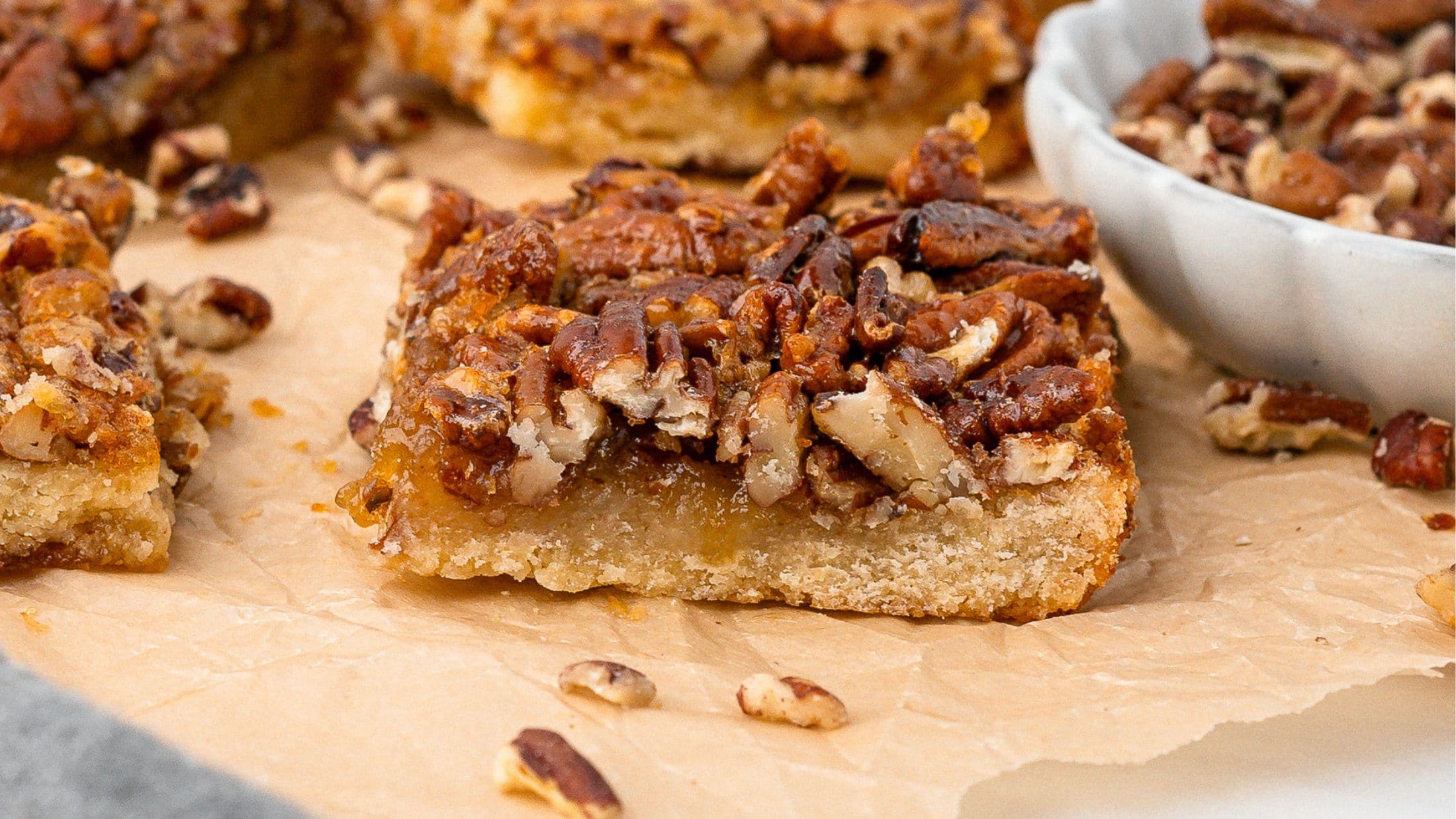 A close up of a pecan pie bar, with chopped pecans around, all on parchment paper