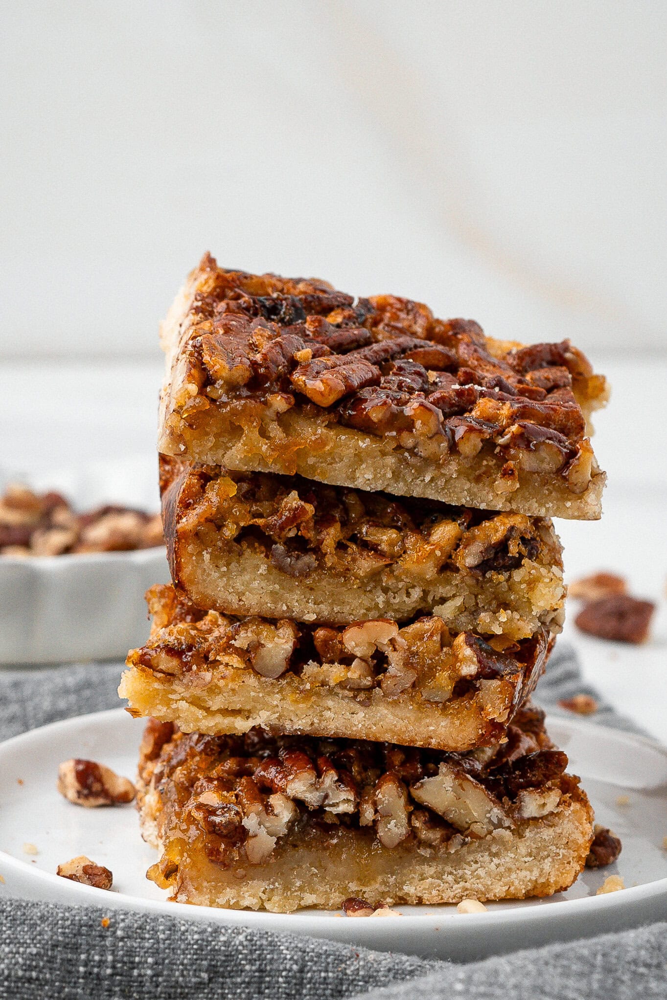 A stack of baked pecan pie bars on a white plate, showing it texture and chopped pecans all around