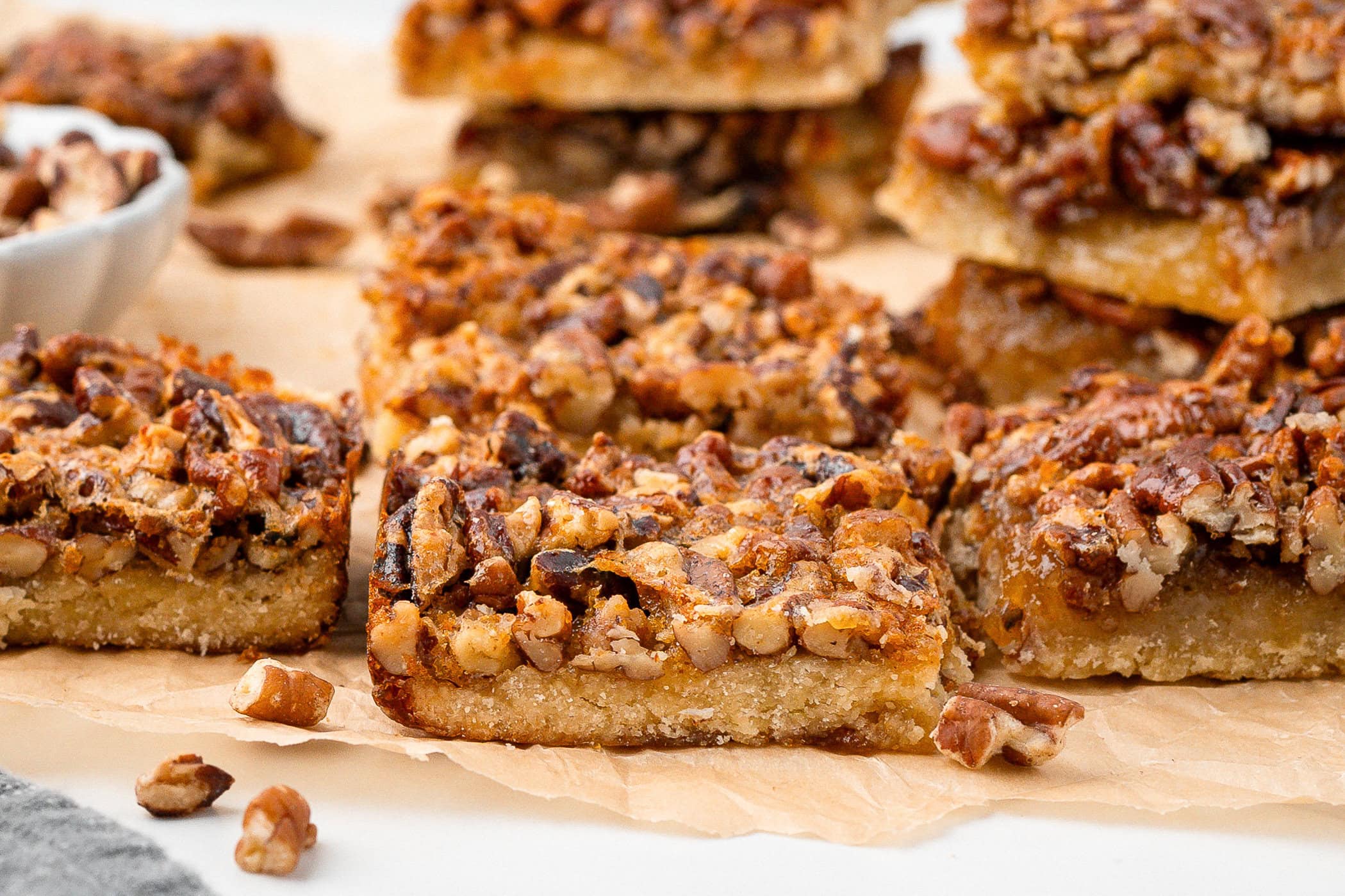 Pecan Pie bars are lined up on parchment paper, chopped pecans all around