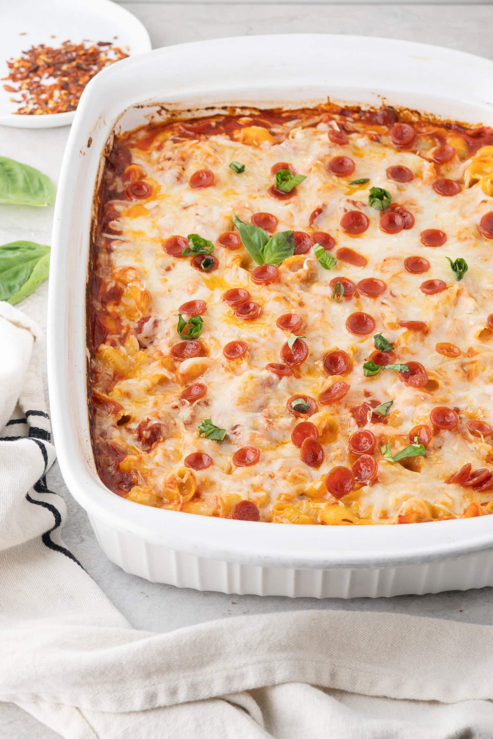 Pepperoni pizza mac and cheese all baked in a baking dish, showing the front part of the pan. Fresh basil and crushed red pepper in the background