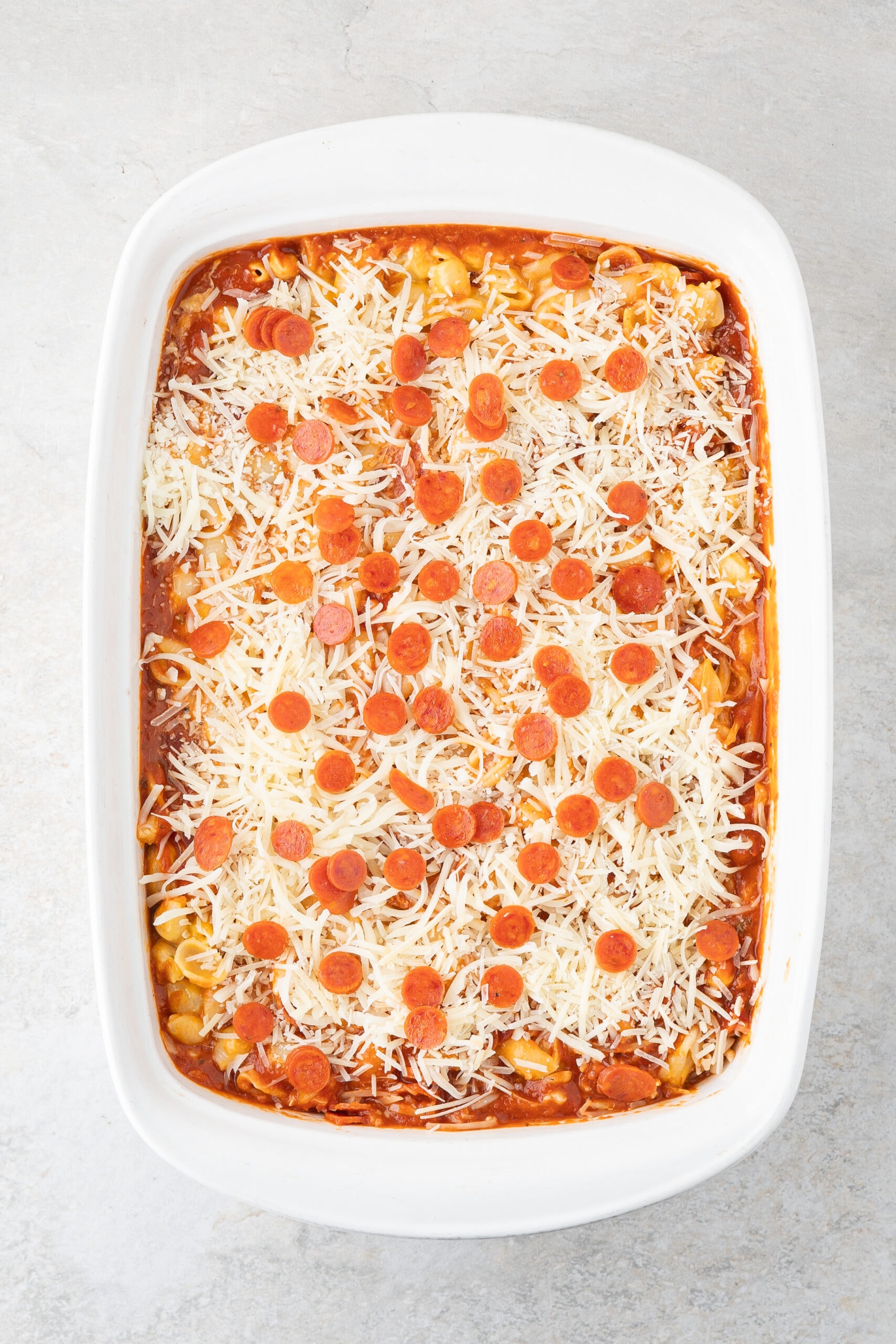 Adding shredded mozzarella cheese and mini pepperoni's on top of pasta and Velvetta cheese in baking dish.