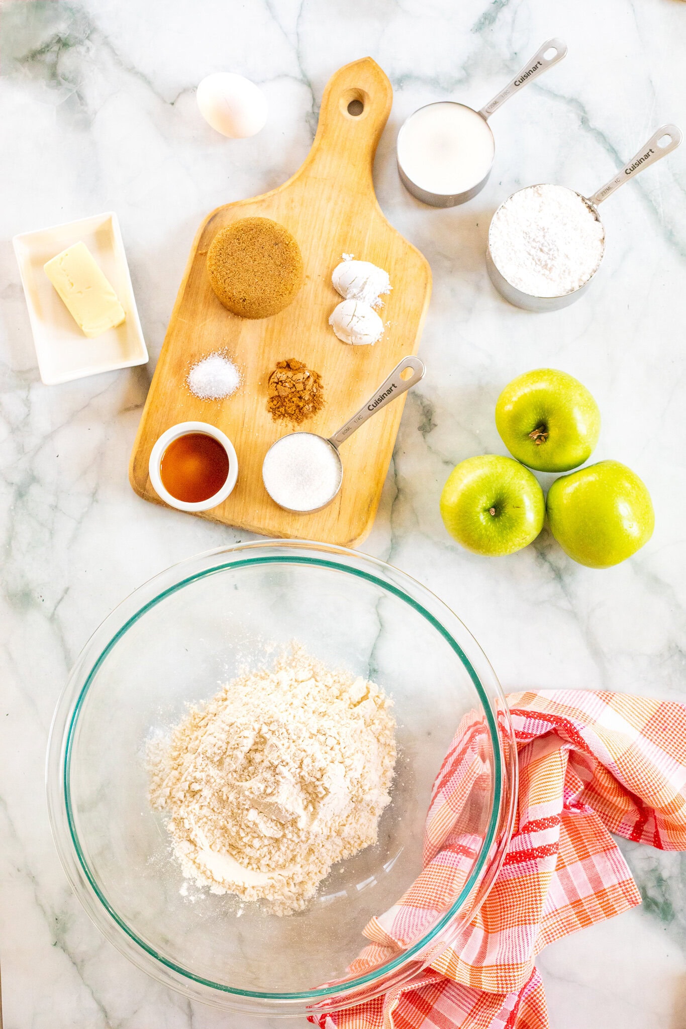 All ingredients to make a caramel apple muffins: brown and white sugar, baking powder, cinnamon, butter, egg, vanilla bean paste, milk, apples, and powdered sugar