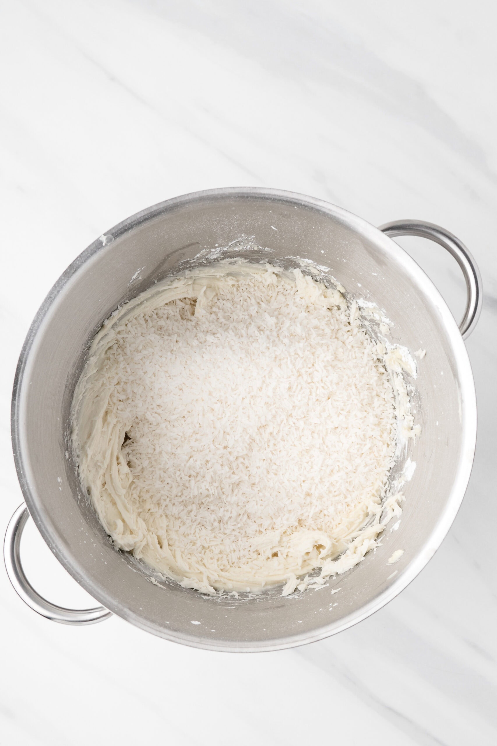 Shredded coconut being added to the mixing bowl of whipped cream cheese and vanilla