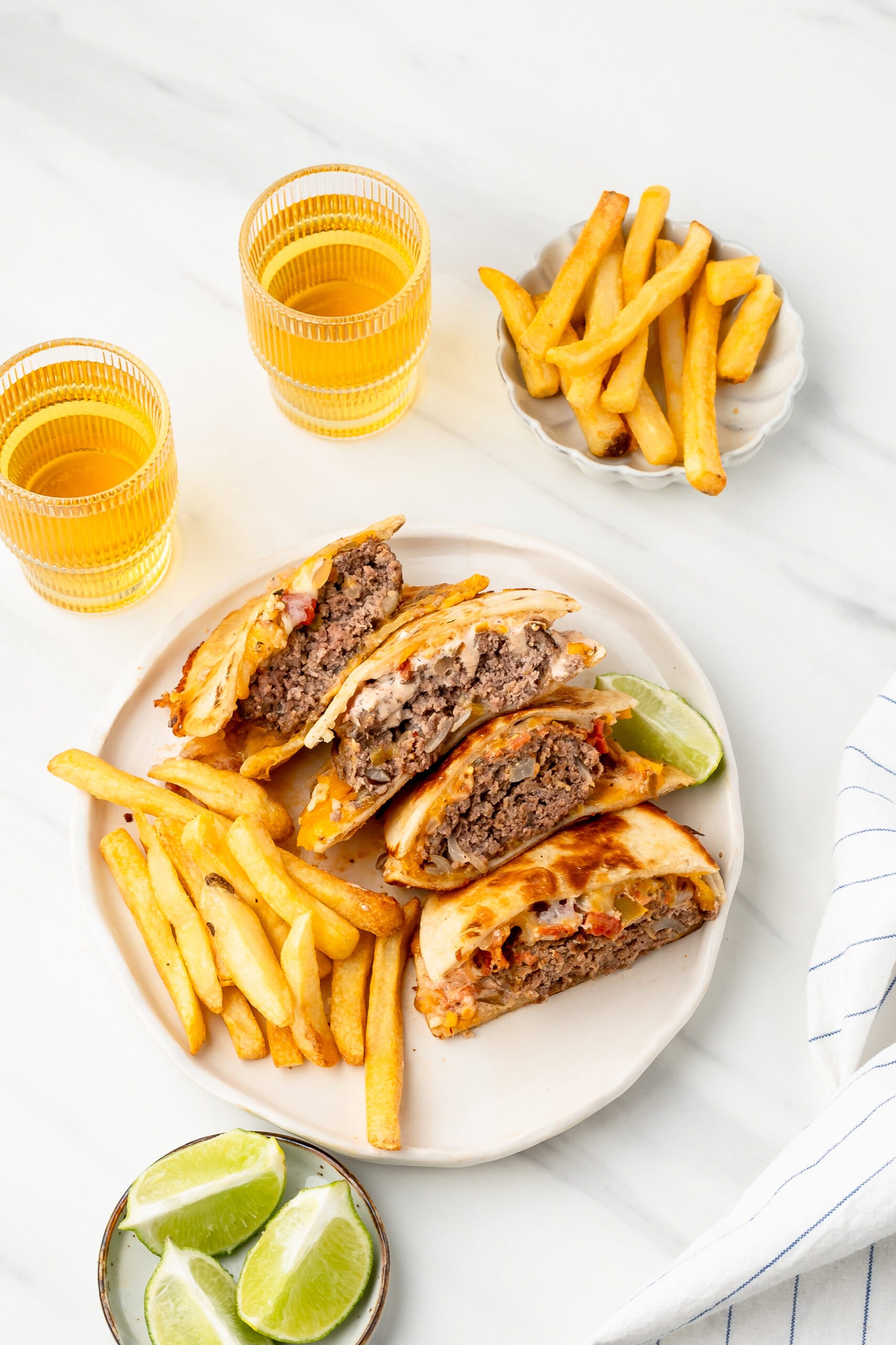 A stack of quesadilla burgers on a plate with fries and lime wedges